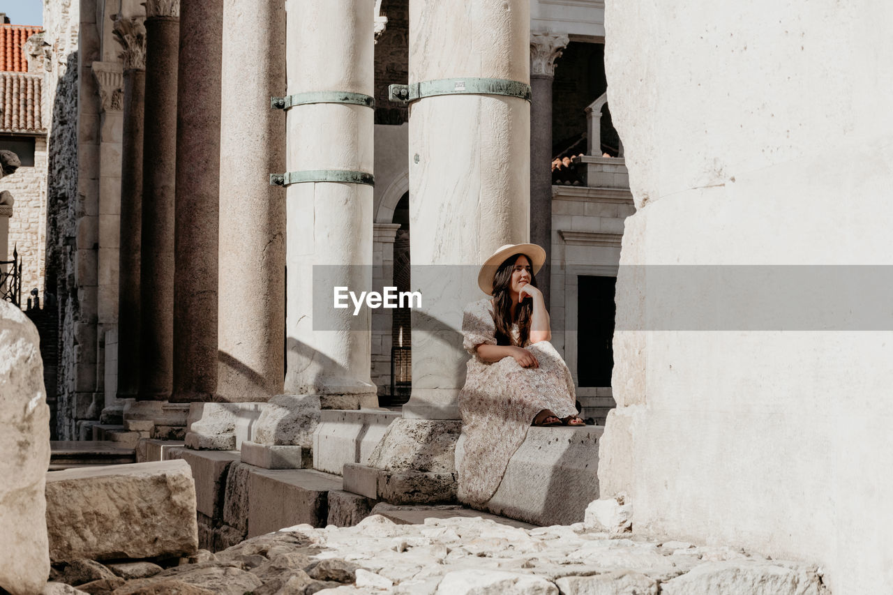 Young woman wearing long dress sitting on stone of ancient ruins in split, croatia.