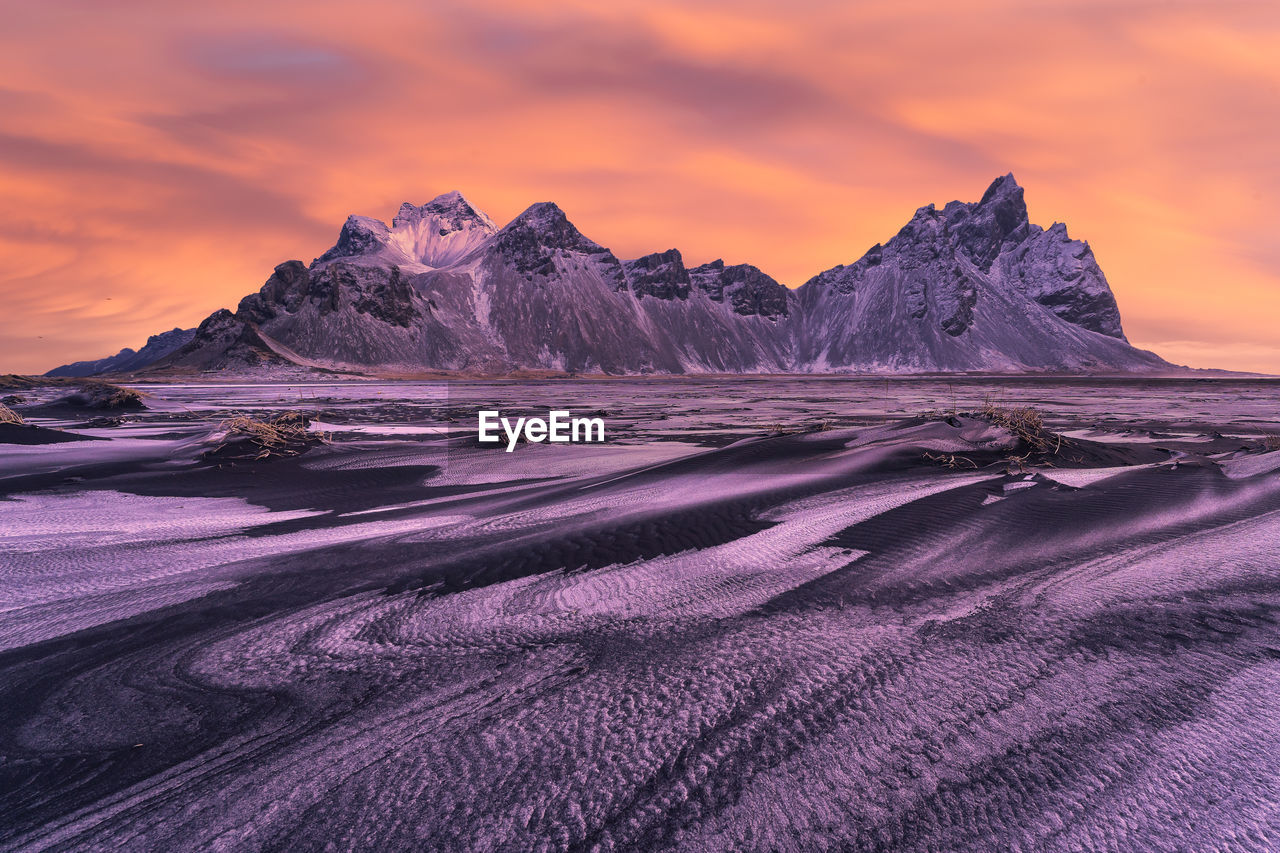 Spectacular nordic scenery of calm frozen lake near rocky vestrahorn mountain with snowy peaks during colorful sunset at stockness beach, iceland