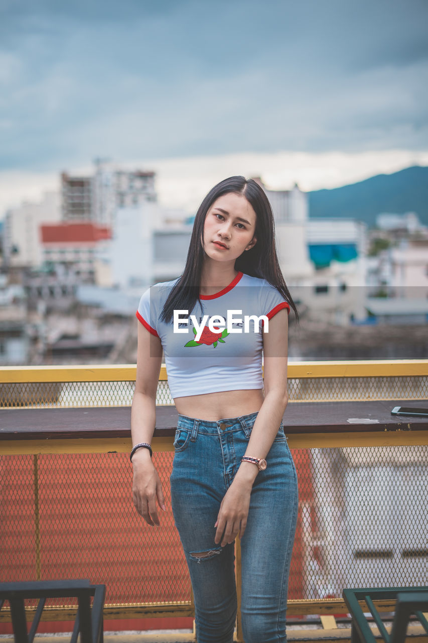 Portrait of young woman standing against cloudy sky in city