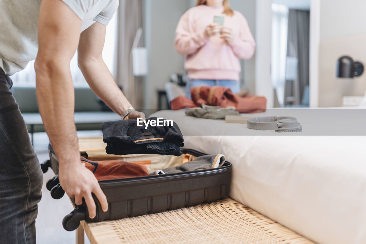 Man removing clothes from suitcase with woman in background at hotel