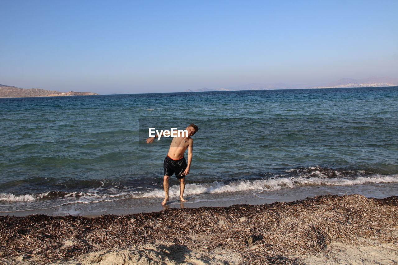 Portrait of shirtless mid adult man standing at beach against clear blue sky