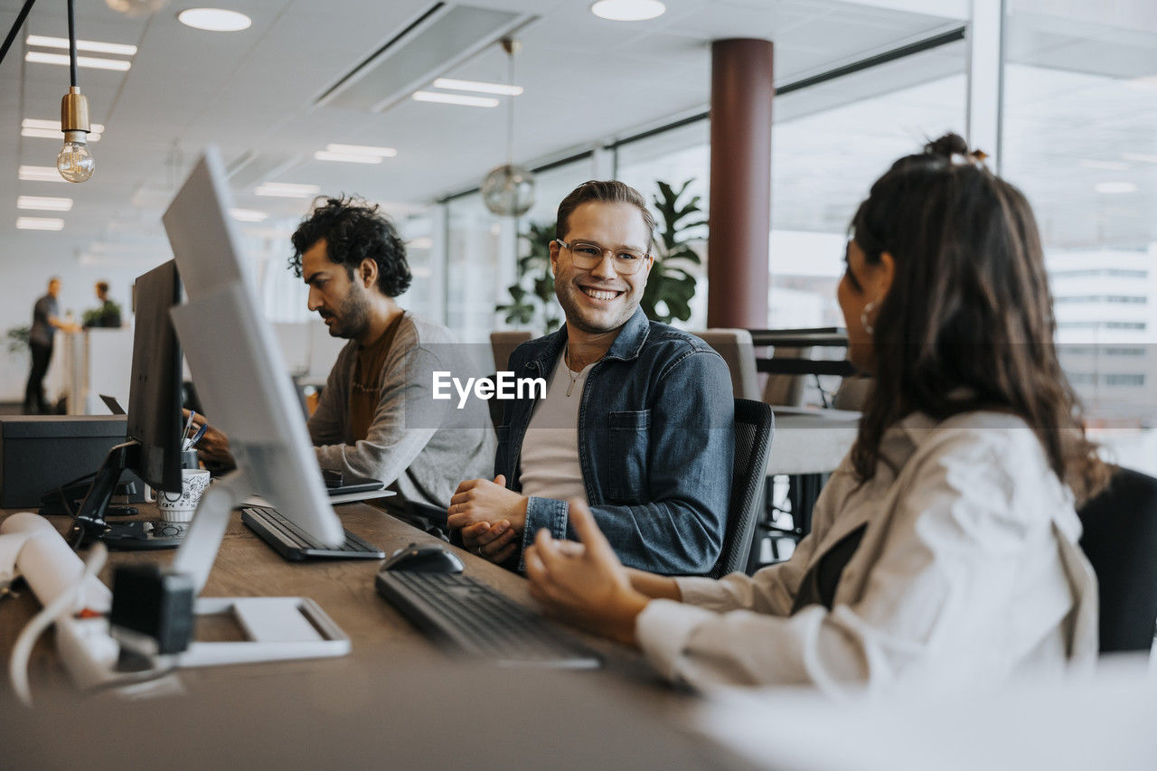 Smiling young male and female colleagues discussing while sitting by businessman at desk in office