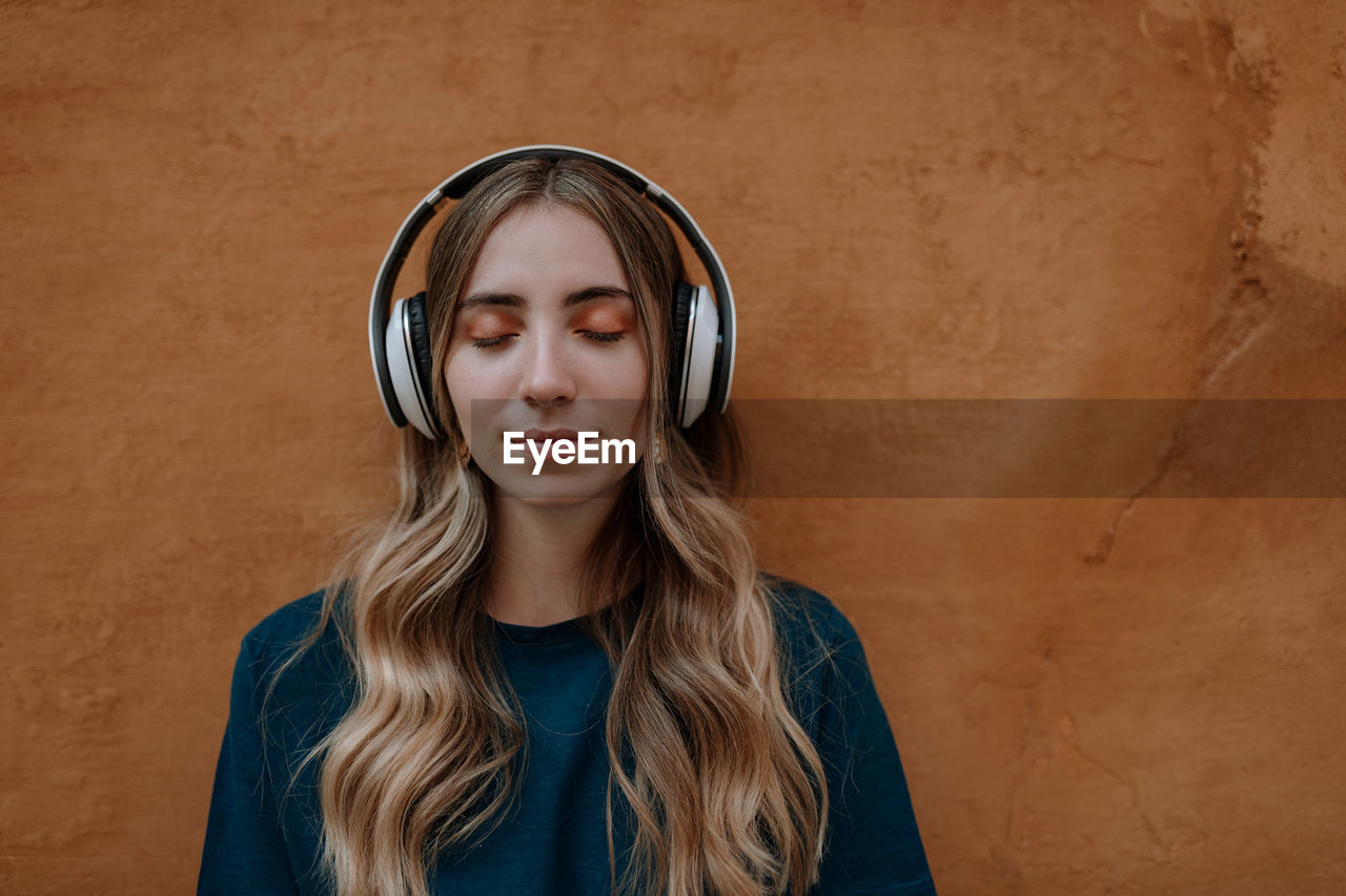 Young mindful blonde female with closed eyes listening to song in wireless headset on brown background in city