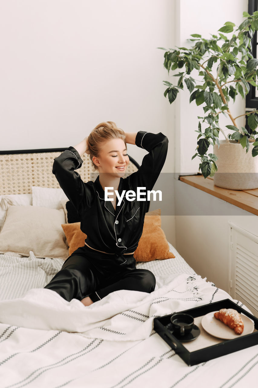 A happy pretty young woman in pajamas is resting and having breakfast in bed in a cozy apartment