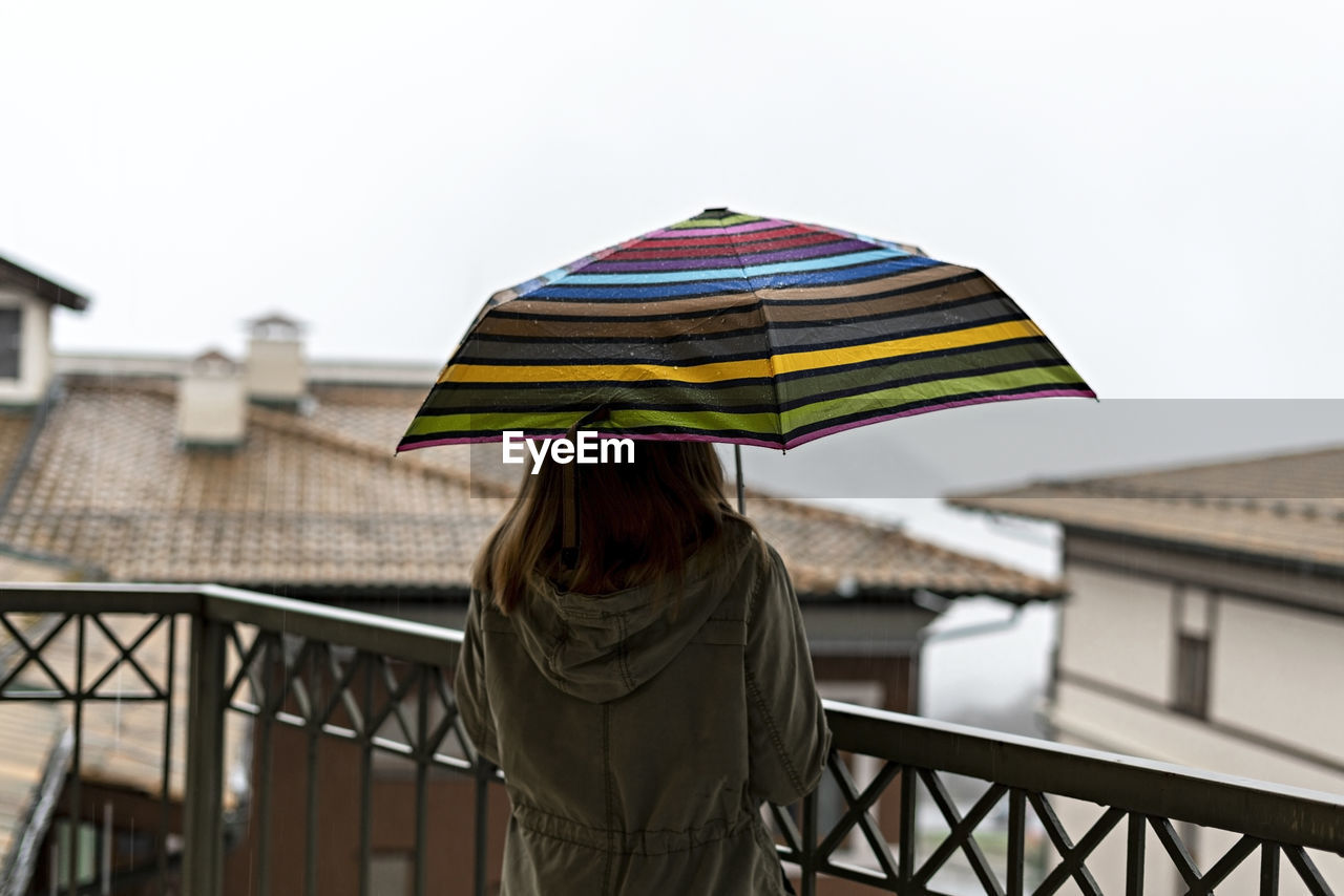Young woman from behind with umbrella in rainbow pattern in drops standing on terrace in rain