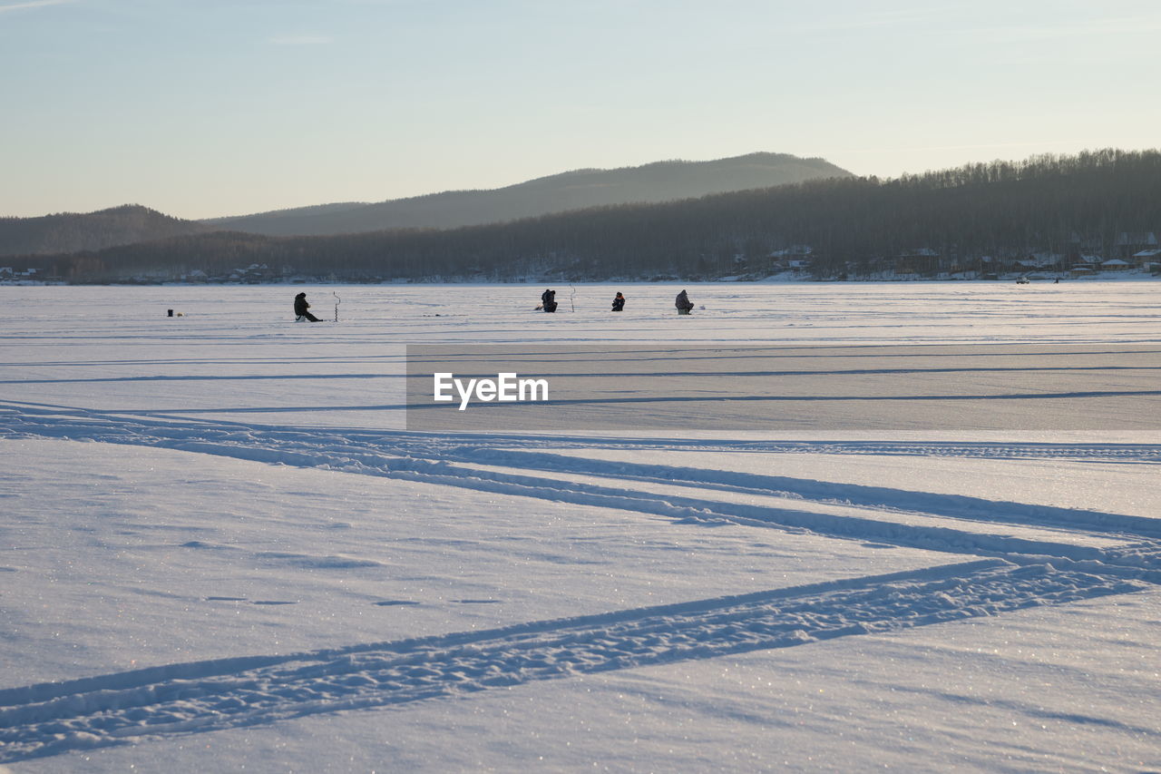 Fishermen sit and fish on a snowy lake, in the evening light.
