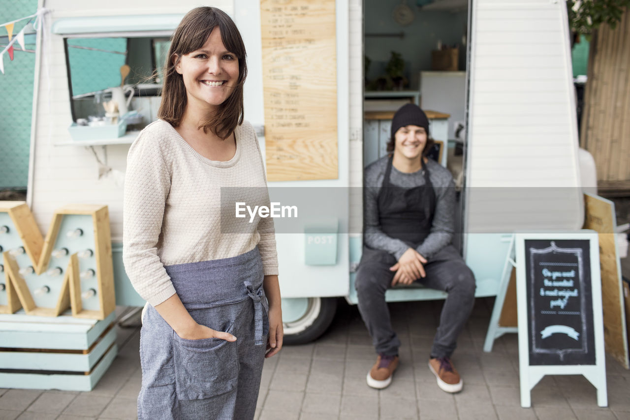 Portrait of happy female owner standing on street while colleague sitting in food truck