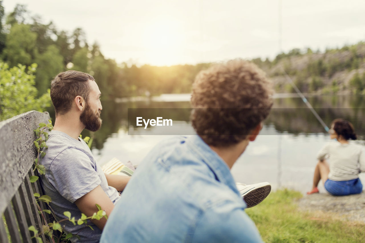 Men sitting on wooden bench while female friend fishing at lakeshore