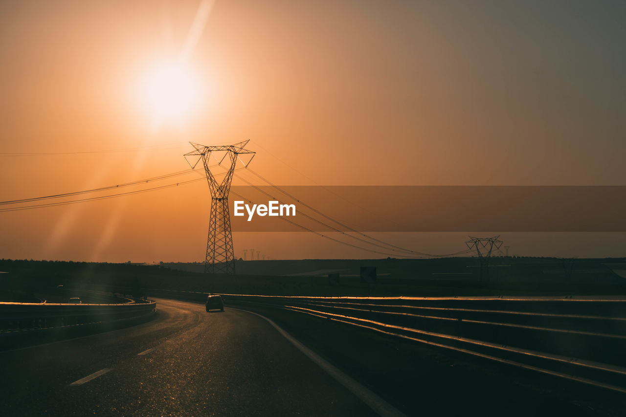 Electricity pylons by road against sky during sunset