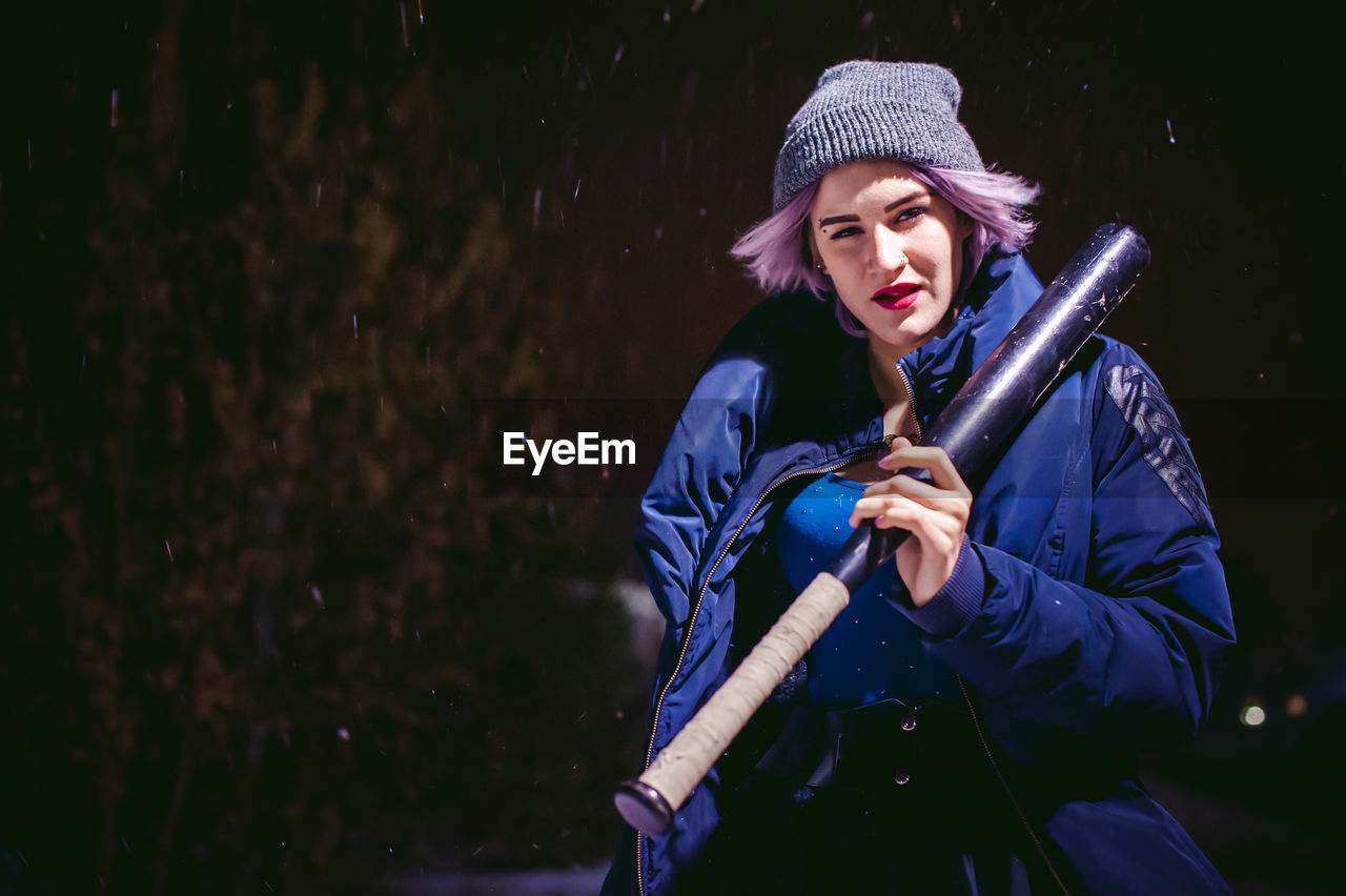 Young woman with baseball bat standing outdoors at night