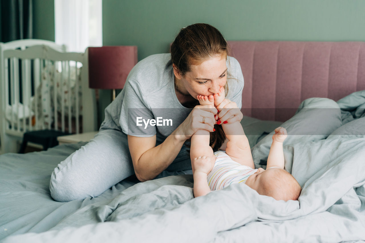 Young happy redhead mom is having fun, kissing the little feet of a newborn baby lying on the bed. 