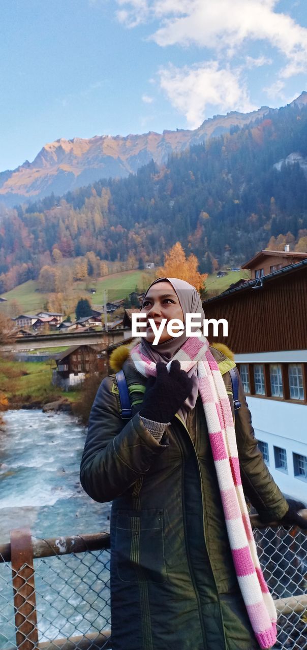 Smiling mature woman looking away while standing on footbridge over river against mountain during winter