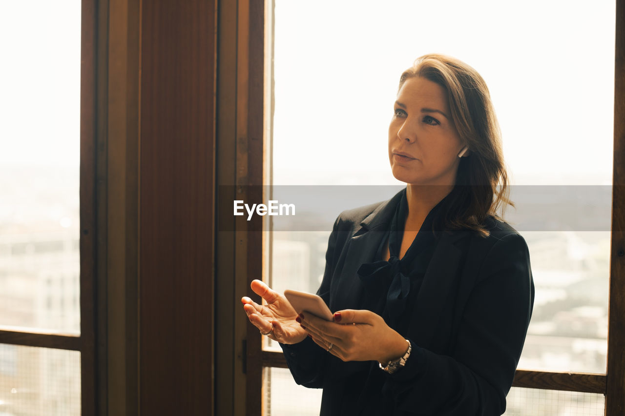 Thoughtful female mid adult lawyer talking on smart phone through bluetooth at office