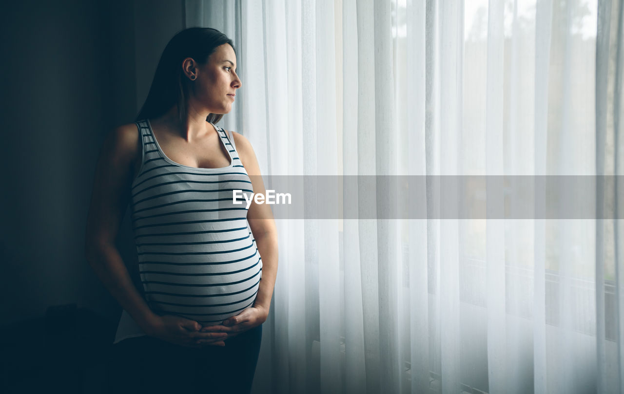 Pregnant young woman looking away while standing against window at home