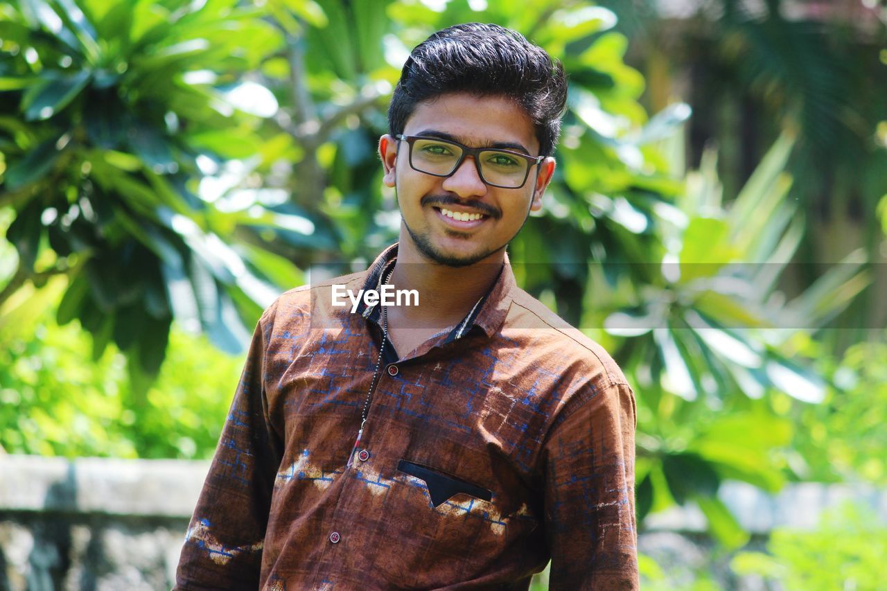 PORTRAIT OF SMILING YOUNG MAN STANDING AGAINST PLANTS