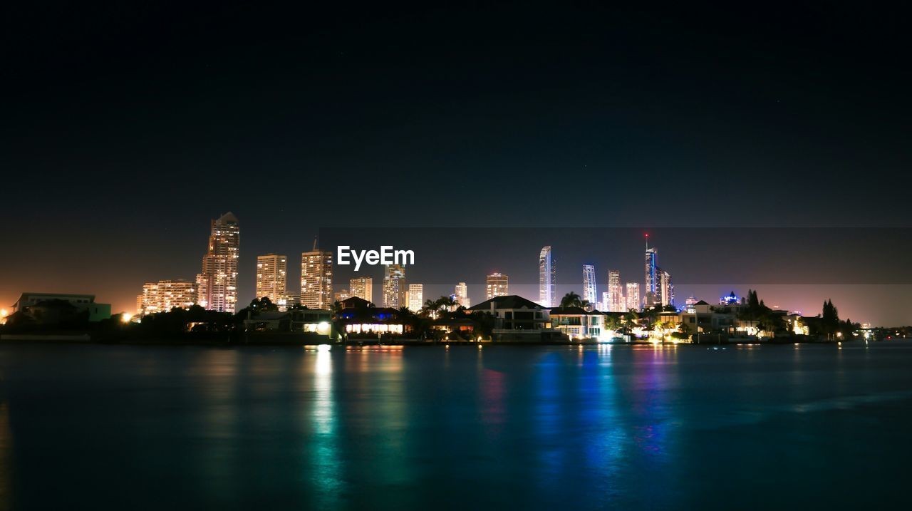 Illuminated modern buildings by lake against sky in city at night