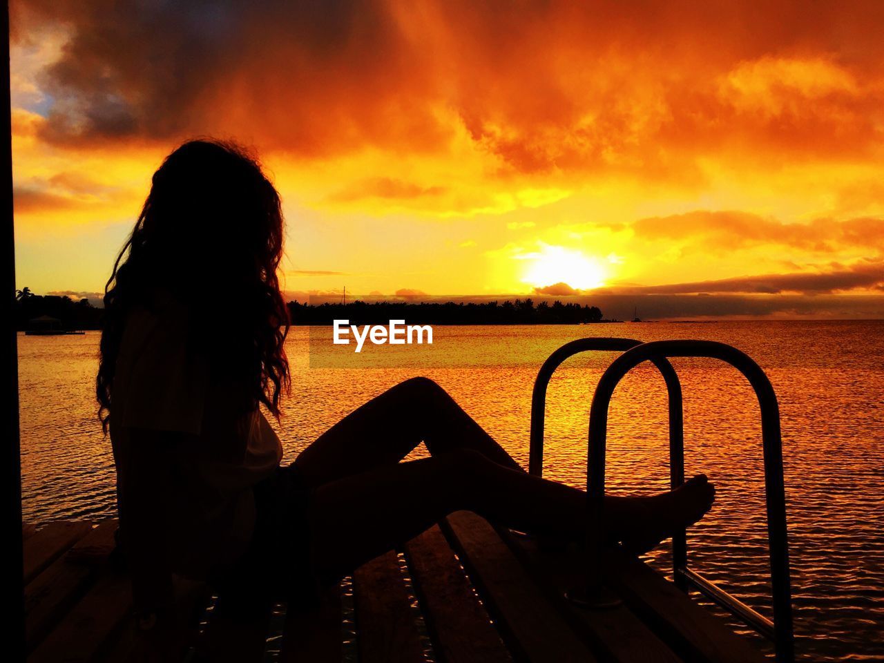 Silhouette woman sitting on pier over sea against orange sky