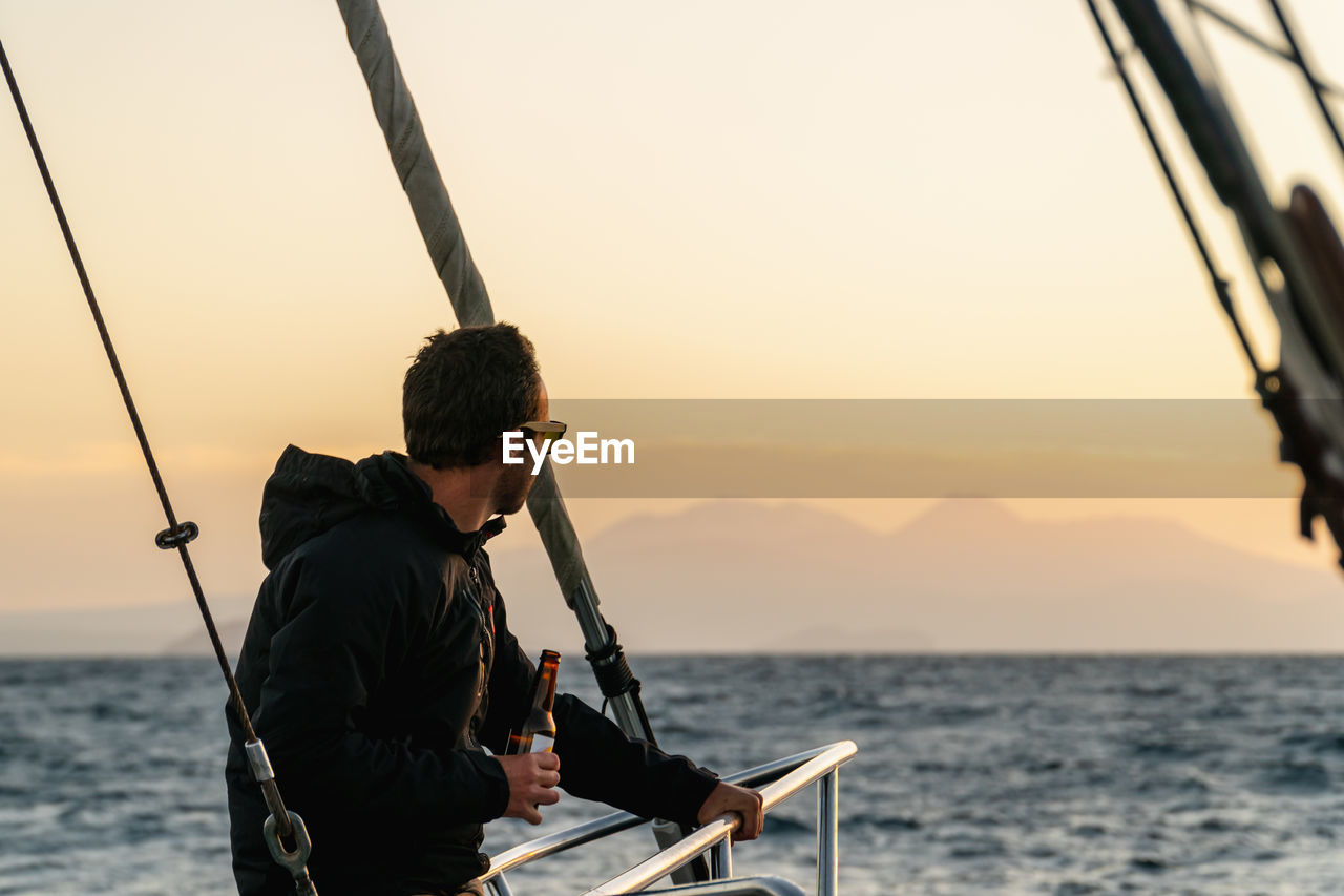 Male on boat bow looking at horizon with beer in hand during sunset at sea on a sailboat.