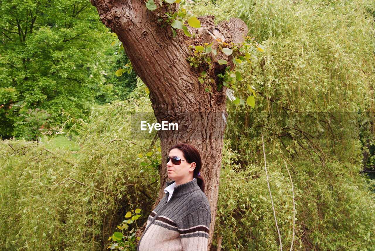 Pregnant woman wearing sunglasses and jacket standing against plants and trees