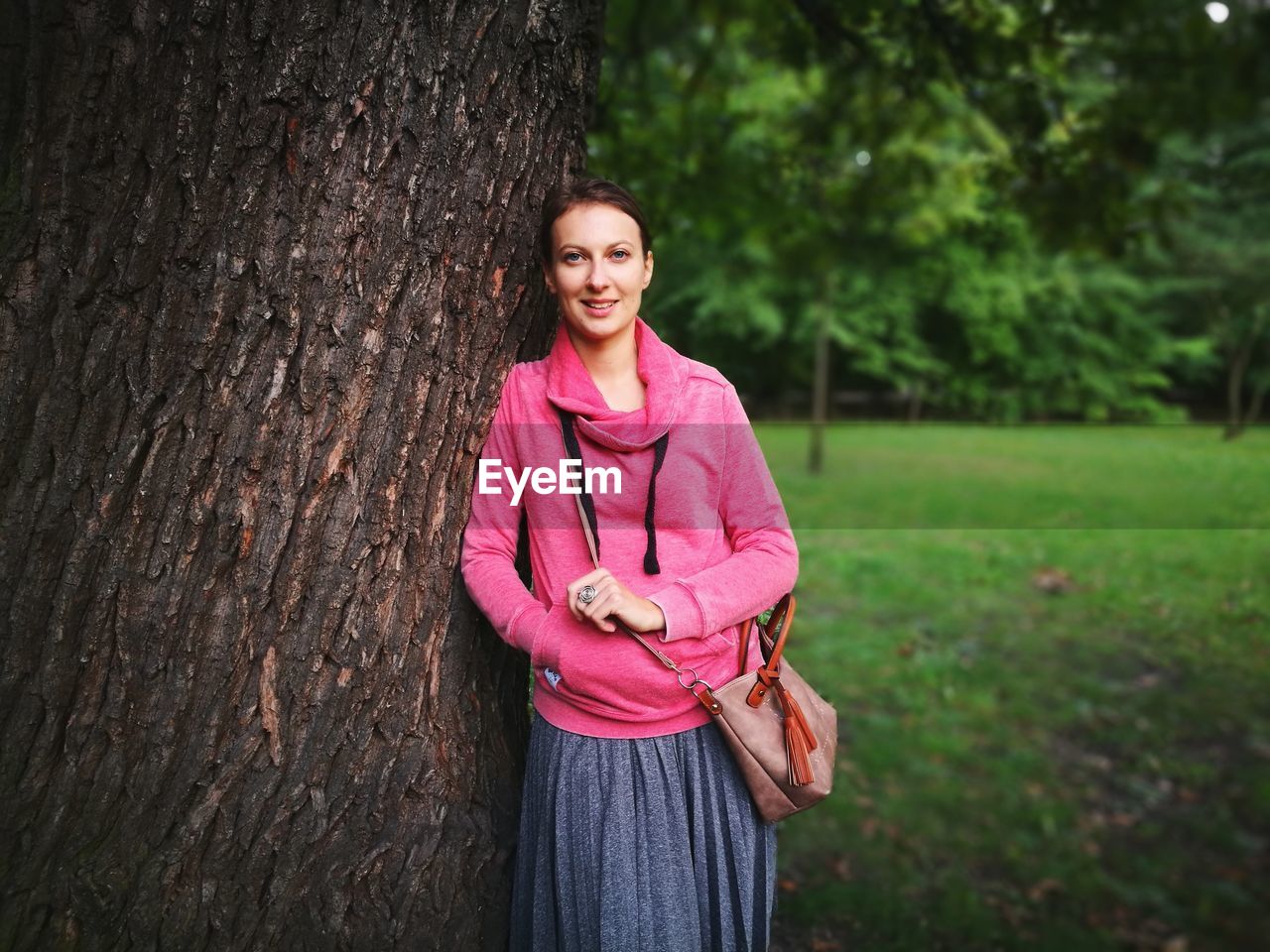 Portrait of smiling young woman standing by tree trunk at park