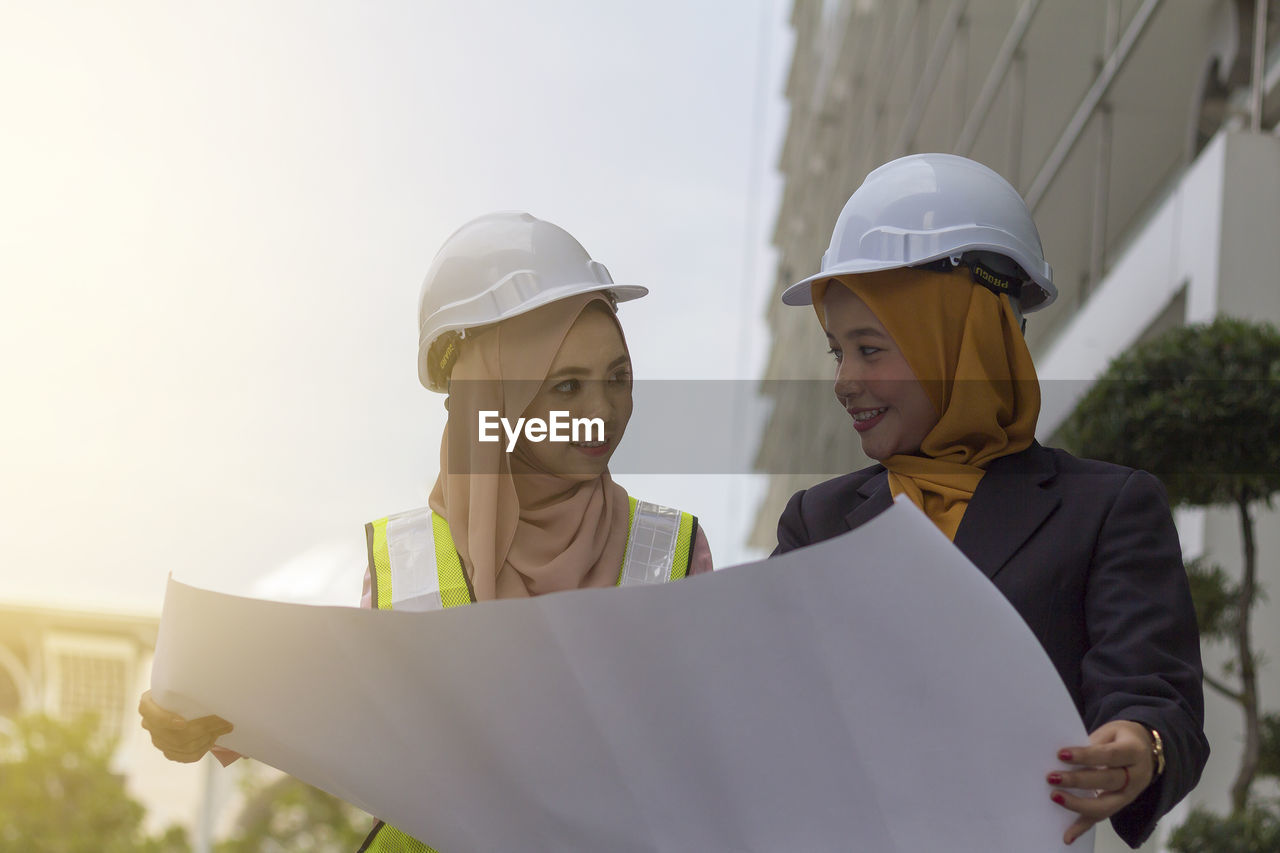 Happy female architects looking at each other while holding blueprint against clear sky