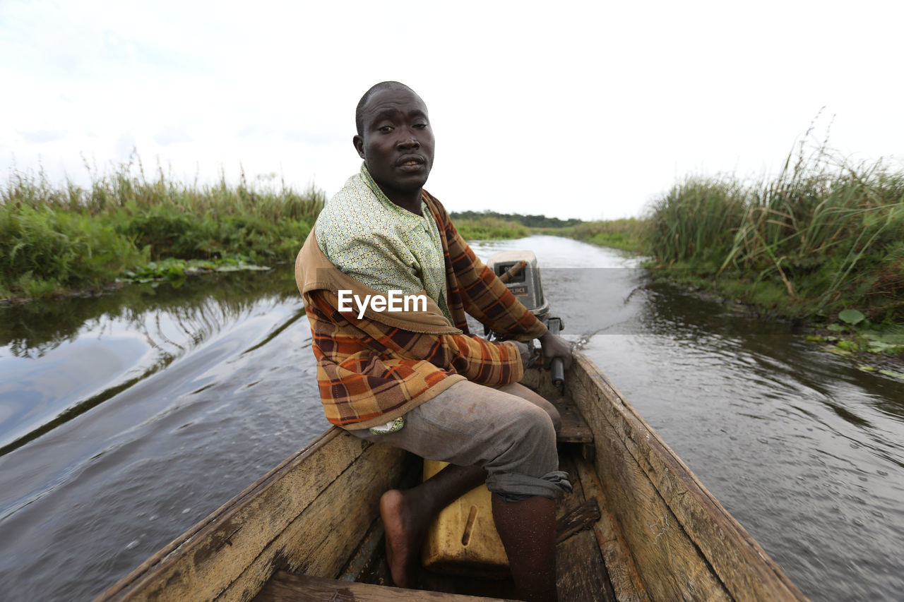 FULL LENGTH PORTRAIT OF YOUNG MAN SITTING ON WOOD AGAINST LAKE