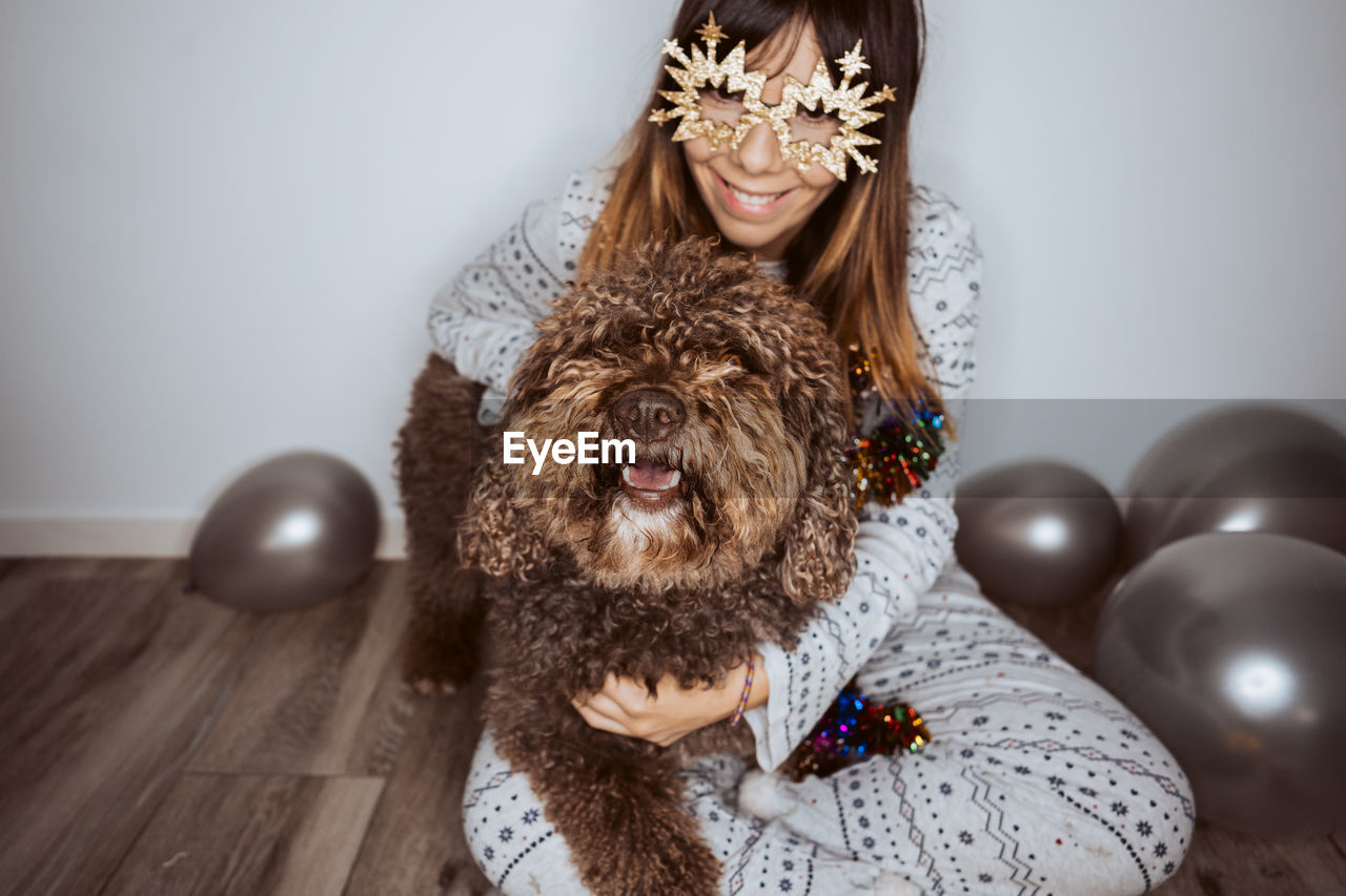 Smiling woman wearing mask sitting with dog and balloons on floor at home