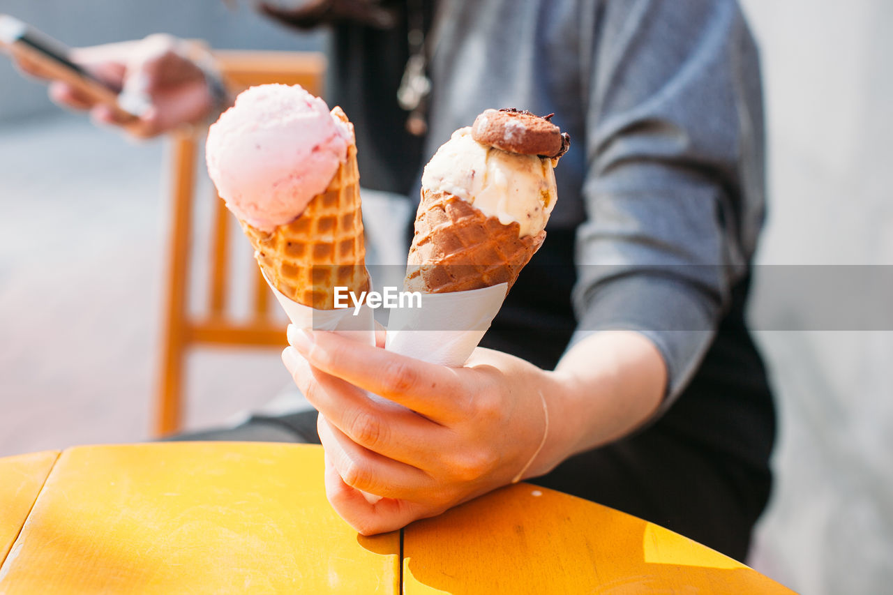 Midsection of woman holding ice creams in waffles while text messaging at restaurant