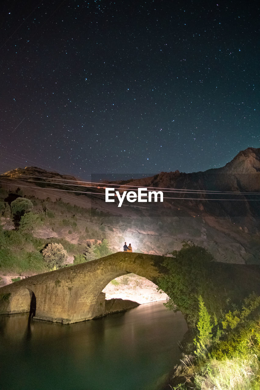 Couple standing on arch bridge over river at night