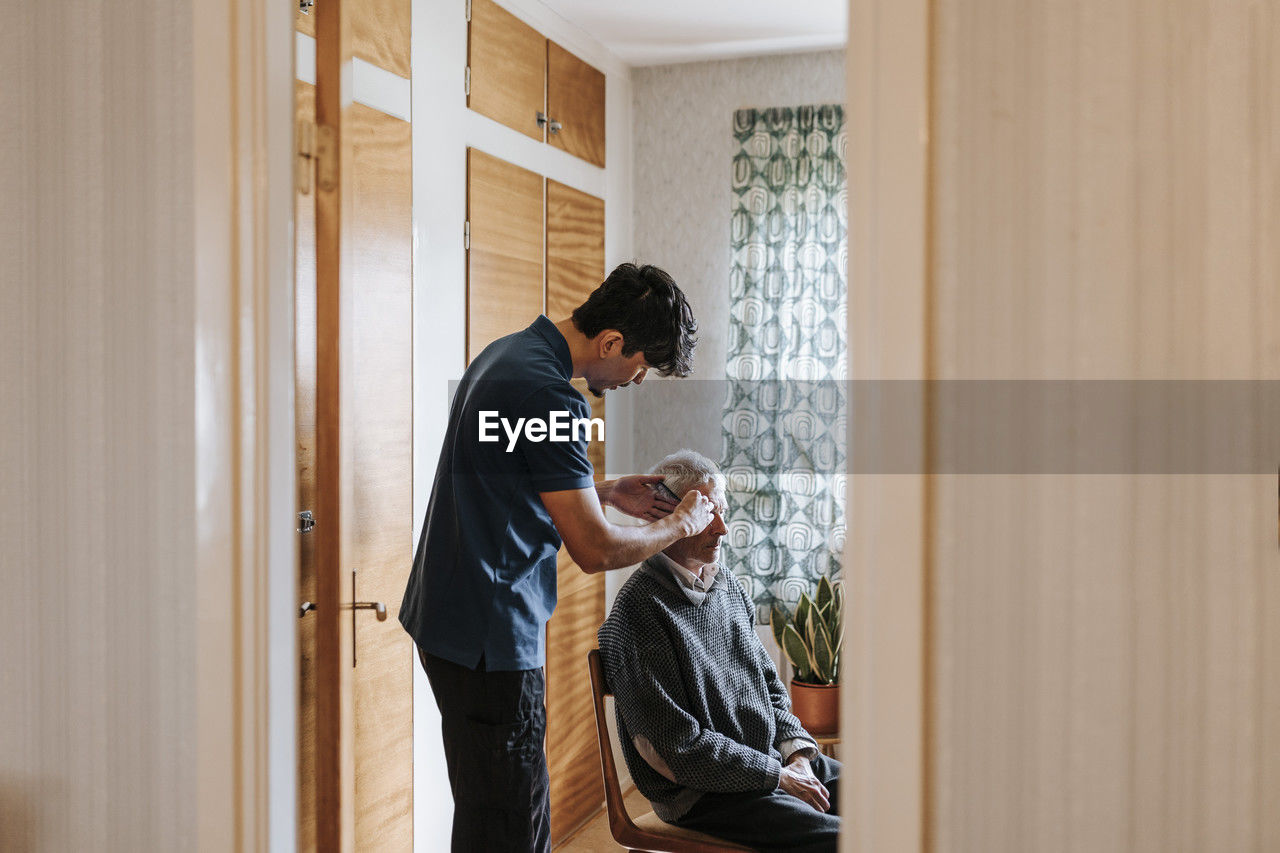 Side view of male care assistant combing hair of senior man sitting at home