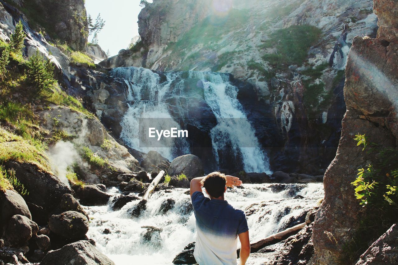 Rear view of man standing against waterfall during sunny day