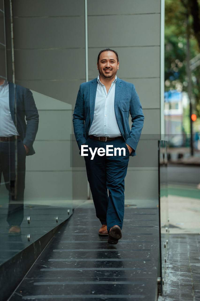 Happy hispanic mature male manager in suit holding hands in pockets and looking at camera with smile while walking near glass wall on sunny day on city street