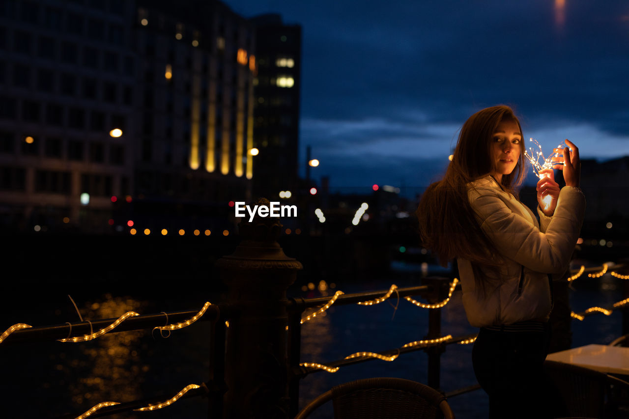 Portrait of young woman holding illuminated string light at night