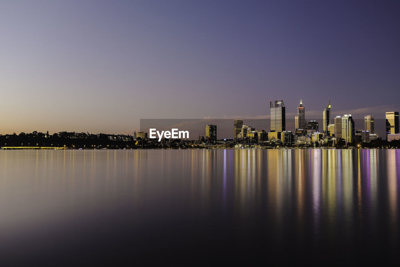 Illuminated buildings by river against clear sky at sunset