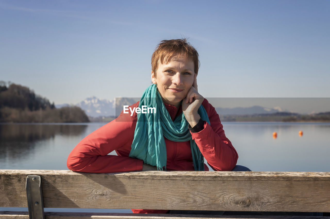 Portrait of woman sitting on bench against lake and sky