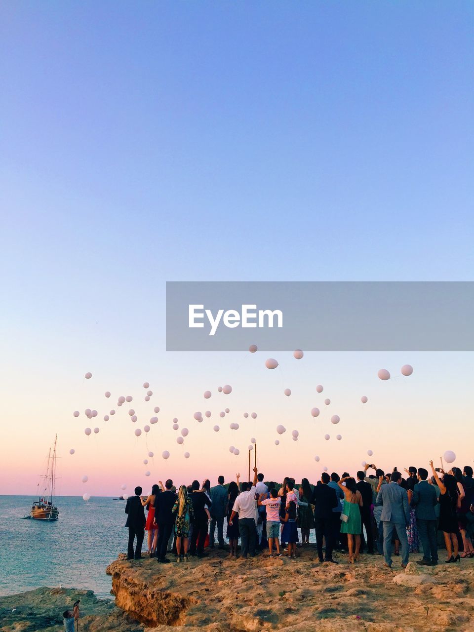 People by sea looking at balloons flying against sky during sunset