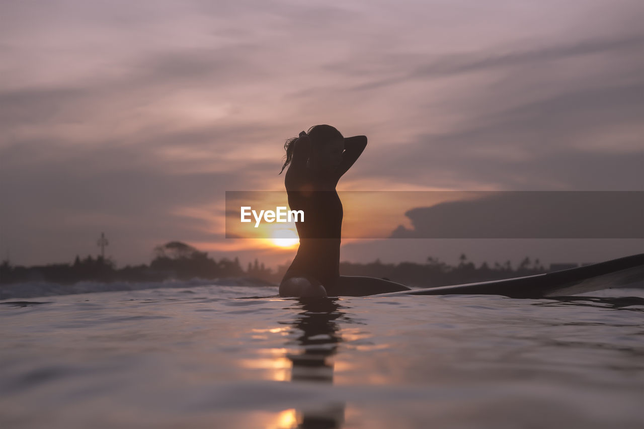 Young woman sitting on surfboard in sea against sky during sunset