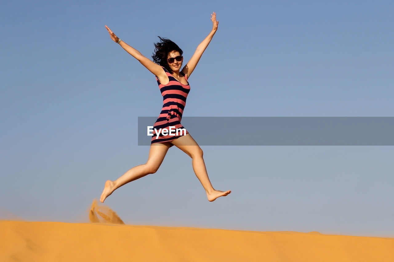 Full length of young woman jumping over sand against clear sky