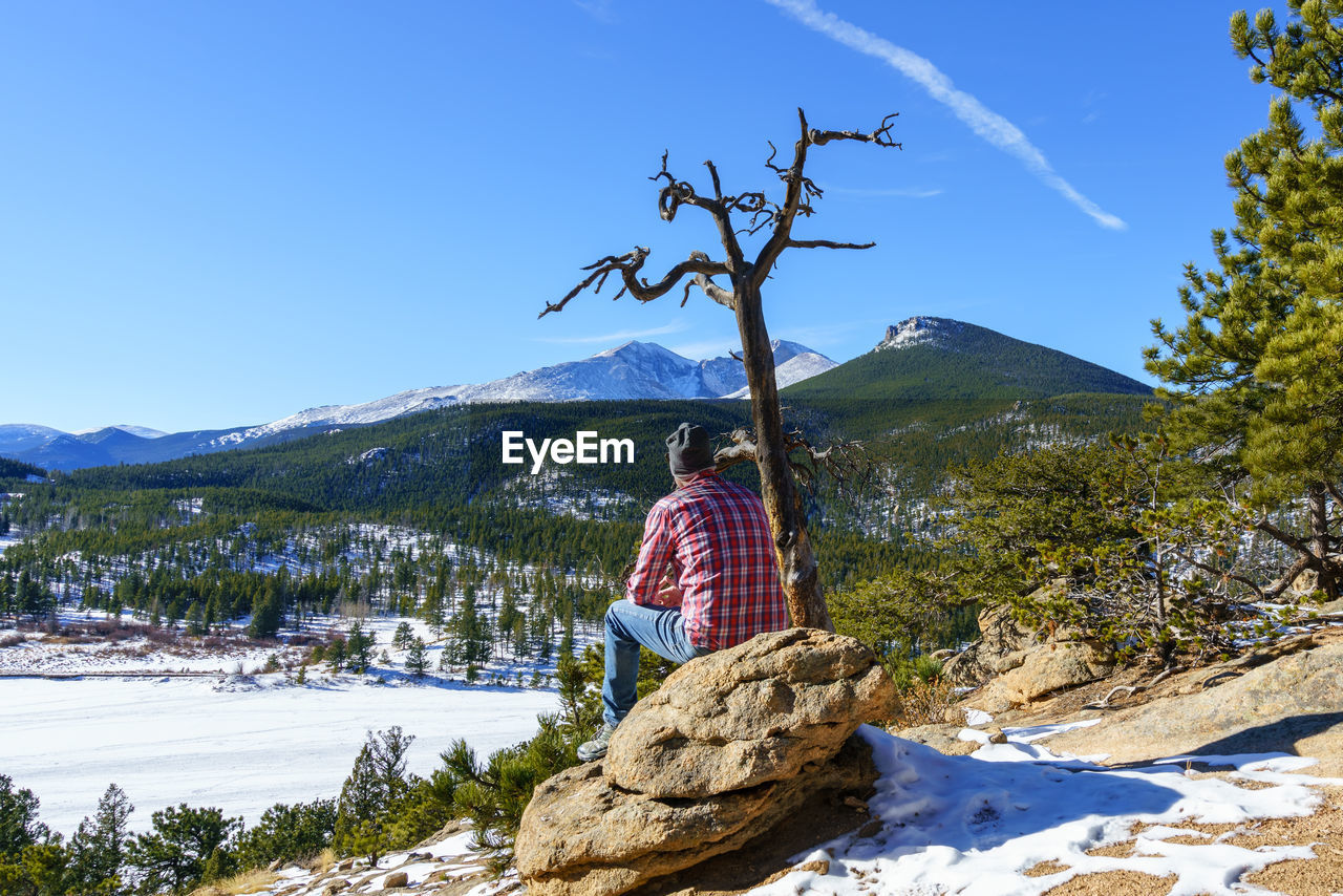 Rear view of man sitting on rock against mountains during winter