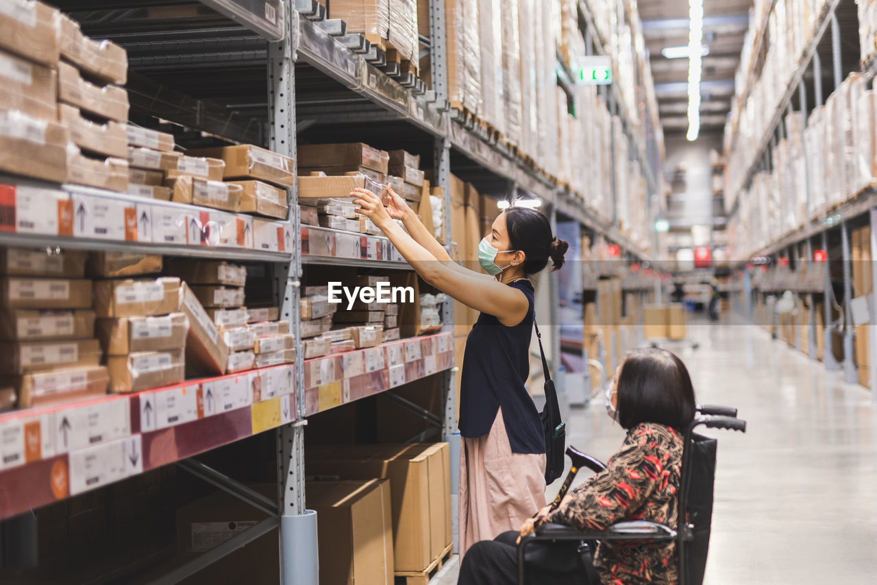 Woman in protective mask hand reaching box in warehouse interior shelves