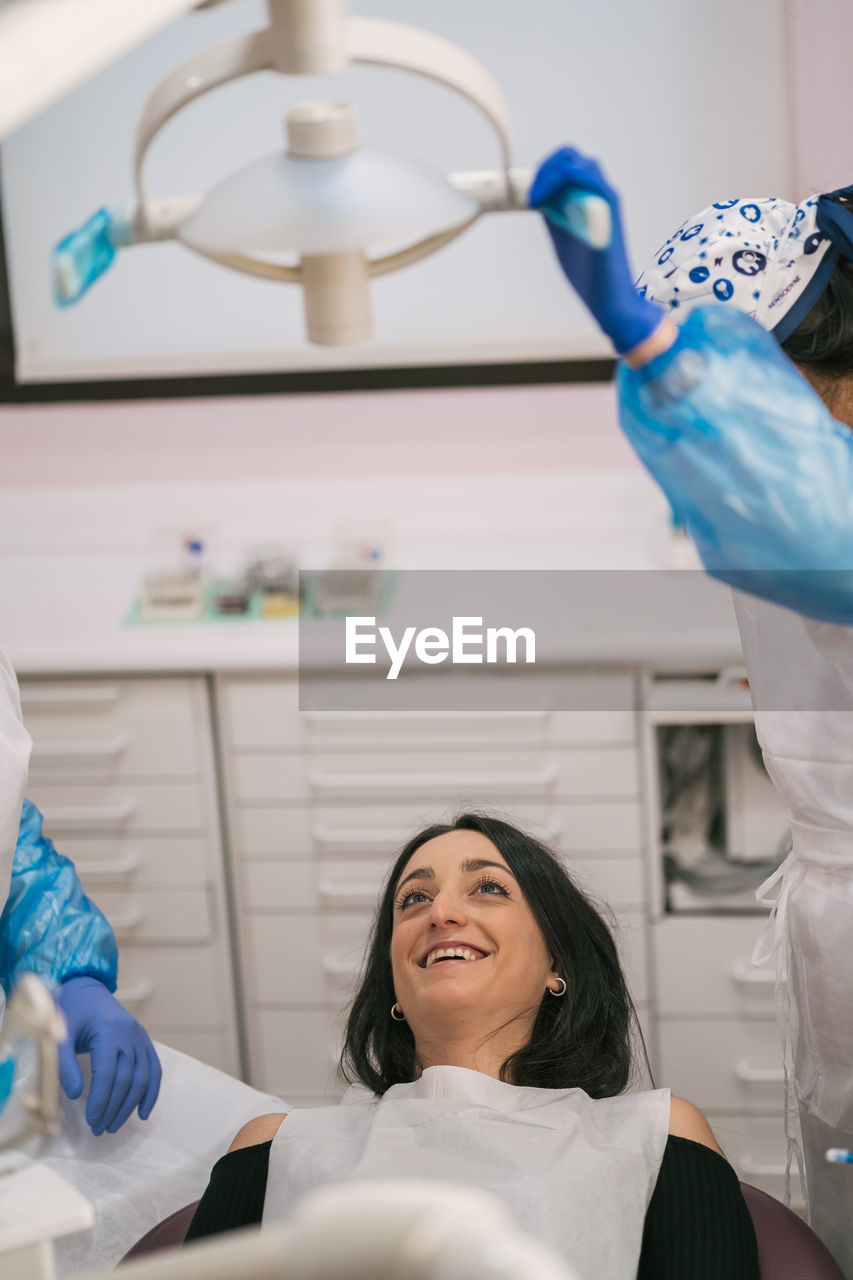 From above of anonymous stomatologist and assistant in uniforms talking to woman lying on medical chair near professional equipment