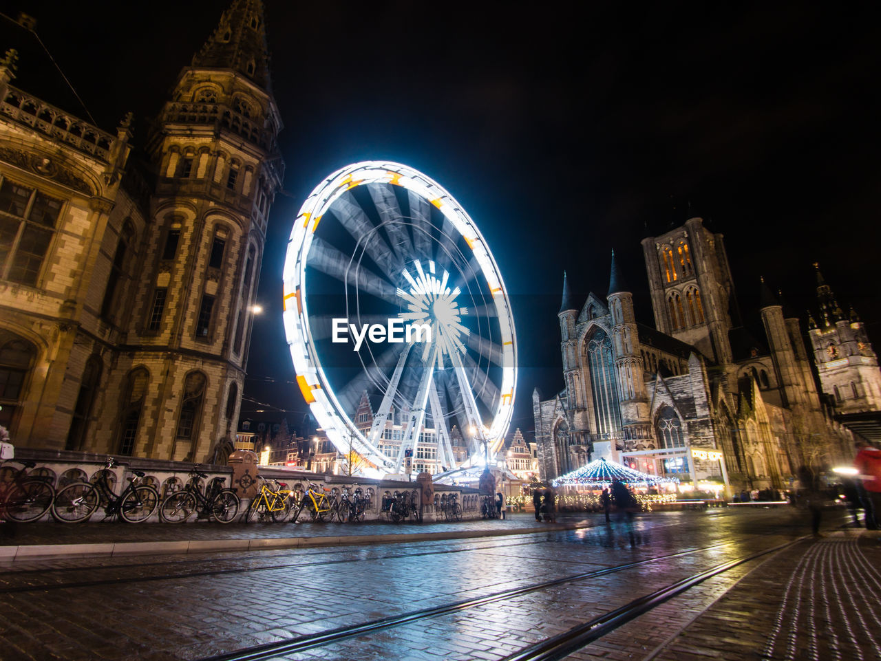 LOW ANGLE VIEW OF ILLUMINATED FERRIS WHEEL AGAINST BUILDINGS AT NIGHT