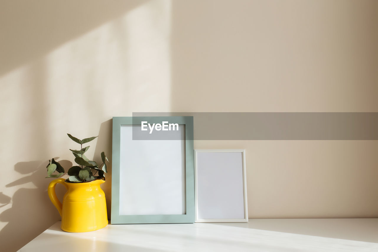 Empty white photo frames on the white table in the interior with beige walls near window.