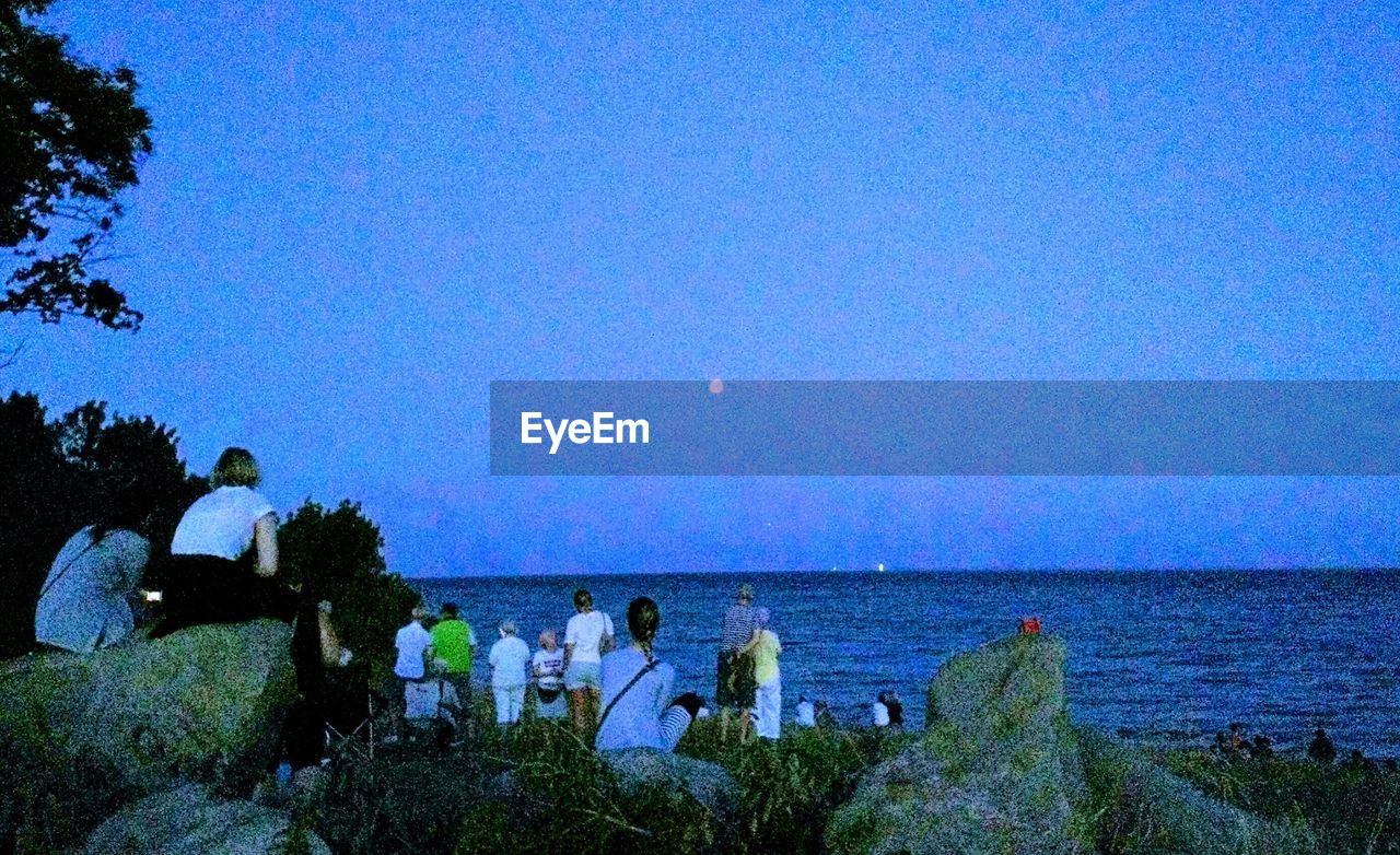 PEOPLE SITTING ON SEA SHORE AGAINST CLEAR SKY