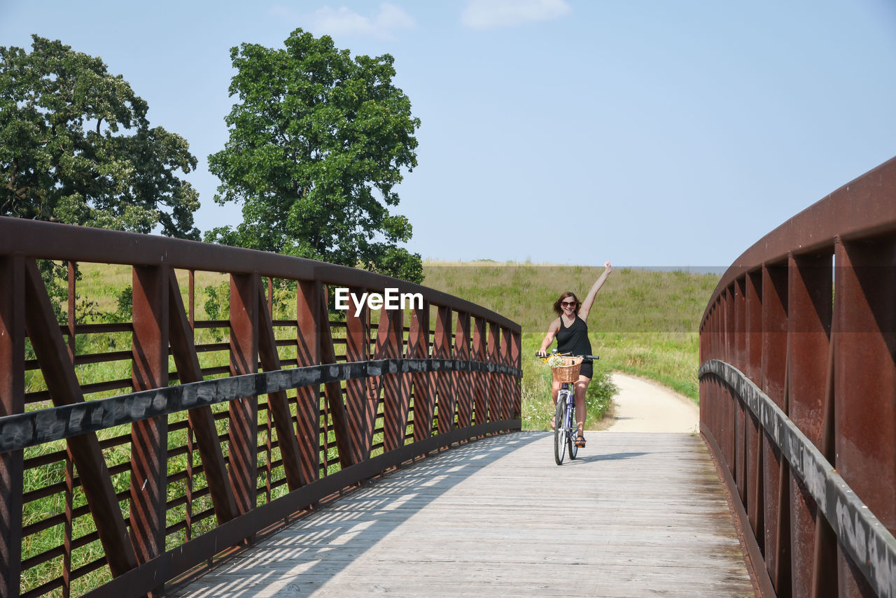 Full length of mature woman riding bicycle on footbridge against blue sky during sunny day