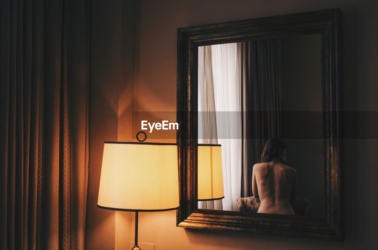 Rear view of shirtless woman reflecting on mirror by illuminated lamp at home