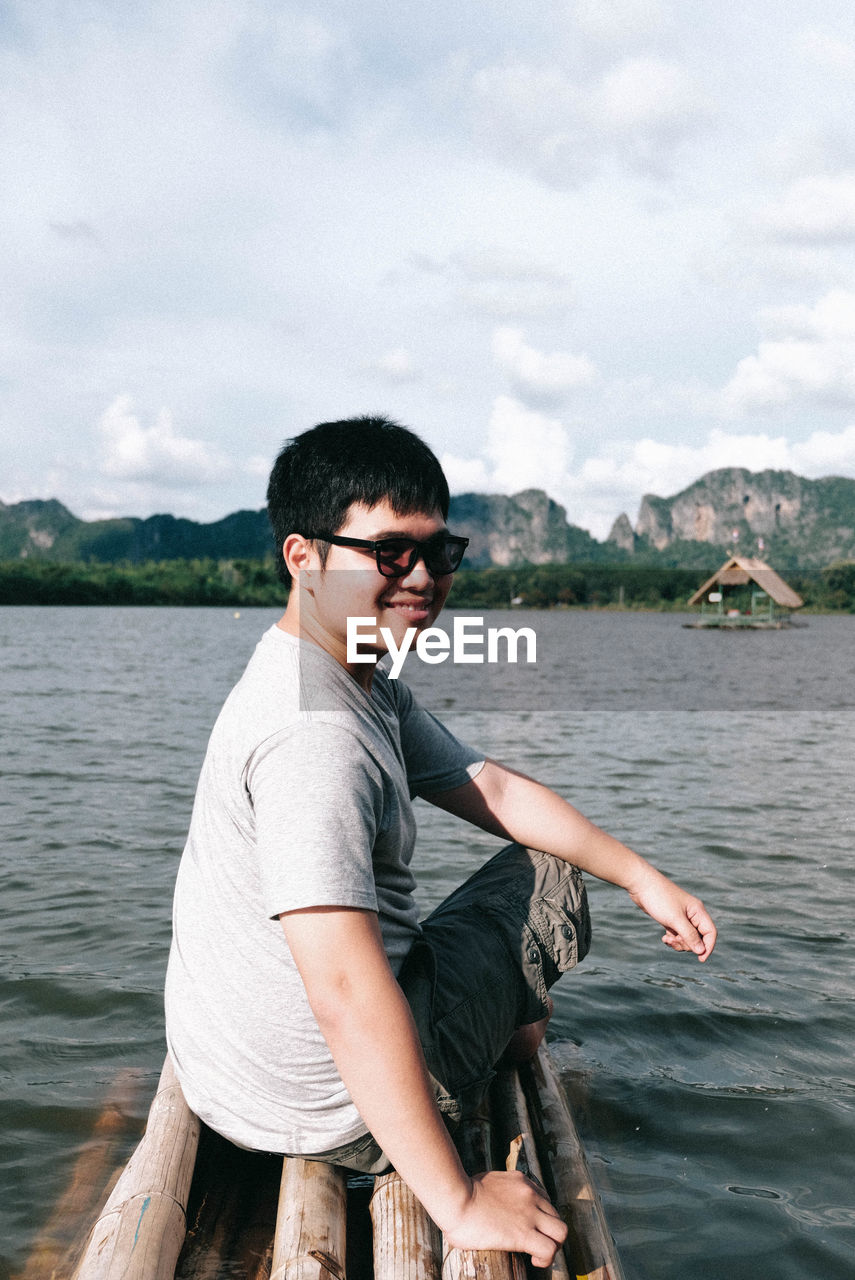 Portrait of man sitting on wooden raft over lake against sky