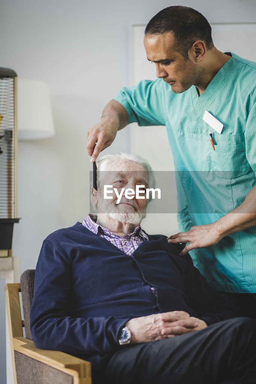 Mature male caregiver combing hair of senior man sitting on chair in nursing home