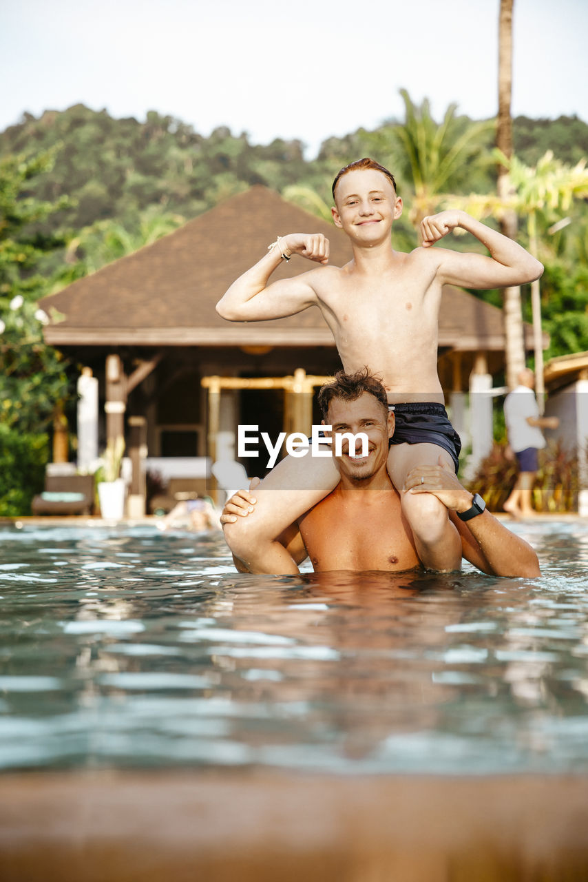 Portrait of father carrying son on shoulders in swimming pool during vacation