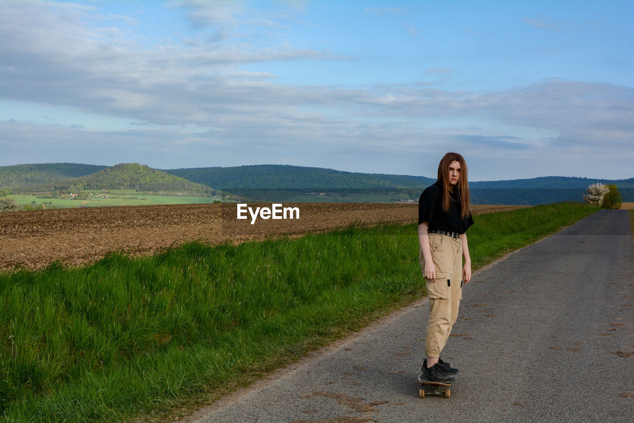 Young girl rides a skateboard on a country road in green nature, with blue sky and many copy space